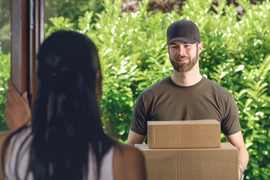 Woman answering the door to an attractive bearded deliveryman carrying two cardboard cartons for delivery view over her shoulder from behind